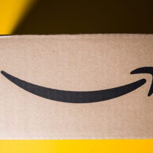A side view of an Amazon delivery box rests against a bright yellow background. The top right corner is covered in shade from the box's shadow.