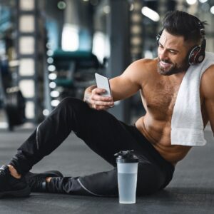 Man sitting on the floor of a gym post-workout holding his phone smiling