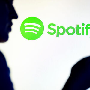 In this photo illustration, a silhouetted individual is seen holding a mobile phone with a Spotify logo displayed in the background.