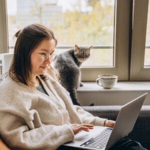 A woman sat on the sofa next to a cat working on her laptop