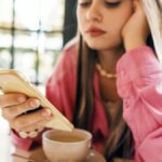 A young woman in a pink shirt checks her phone, with a cup of coffee in front of her