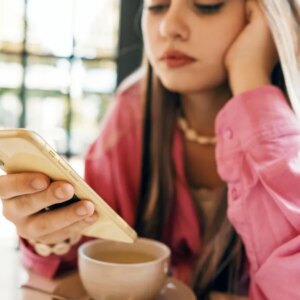 A young woman in a pink shirt checks her phone, with a cup of coffee in front of her