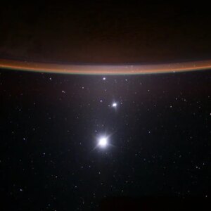 Earth's crescent looks like an orangey sliver with a backdrop of space below it. The moon shines as a bright ball of light while Jupiter and Venus are arranged as bright dots above it.