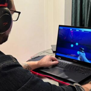 A man wears over-ear headphones while sitting at a desk with hands on a laptop and mouse.