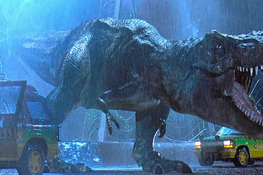 A still from the 1993 Jurassic Park movie. It shows a T. rex with gaping jaws and huge teeth lumbering across a road on a stormy night, between two park vehicles.
