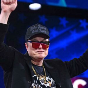 Elon Musk at an event with his hands raised in the air, wearing a cap and reflective sunglasses