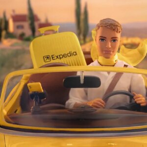 ken doll drives yellow car with expedia suitcase.