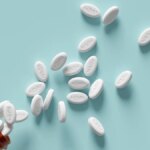 White pills labelled NAD+ spilling out of a brown pill bottle onto a light blue surface.