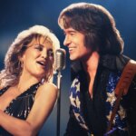 A blonde woman sings and plays a tamborine next to a man that looks like Neil Diamond.